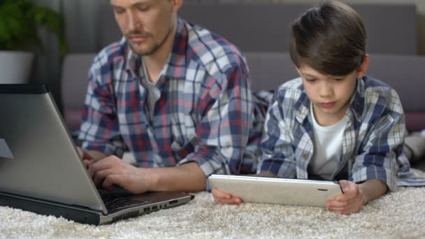 Small son playing on tablet on the floor at home while dad working on laptop — Stock Video