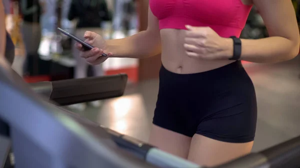 Занятая молодая женщина с помощью гаджета во время тренировки в тренажерном зале, упражнения на беговой дорожке — стоковое фото