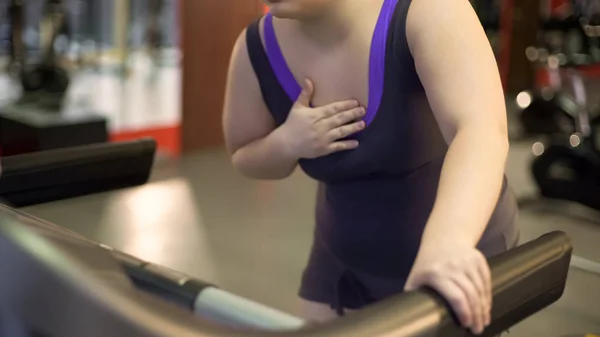Senhora com excesso de peso sofrendo de dor cardíaca e dificuldade em respirar, treino em esteira — Fotografia de Stock