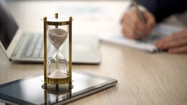 Reloj de arena de pie sobre la mesa con goteo de arena, el hombre que trabaja con gráficos, datos — Foto de Stock