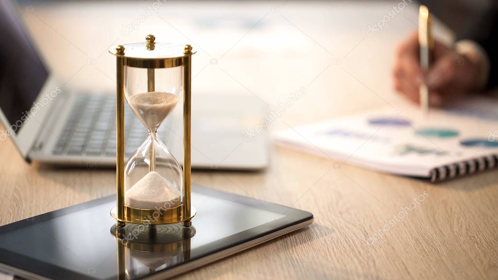 Time is running in hourglass, female manager checking graphs at office desk