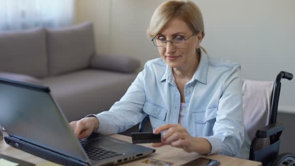 Mujer adulta feliz en silla de ruedas introduciendo el número de tarjeta en el ordenador portátil, compras en línea — Vídeo de stock