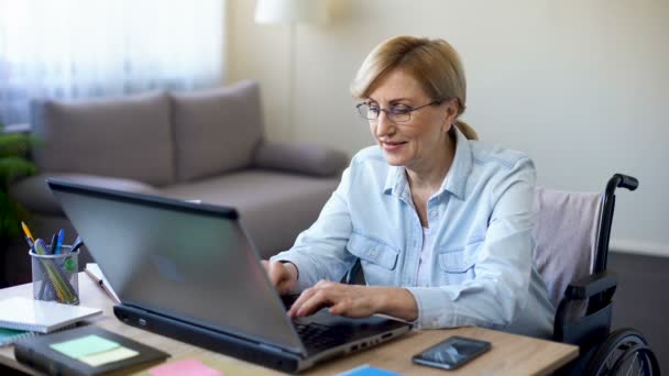 Mulher sênior bem sucedida em cadeira de rodas trabalhando no laptop, conversando com o cliente — Vídeo de Stock