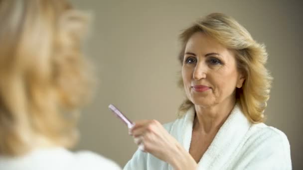 Atractiva mujer mayor cepillando el cabello en el espejo delantero y sonriendo, belleza envejecida — Vídeos de Stock