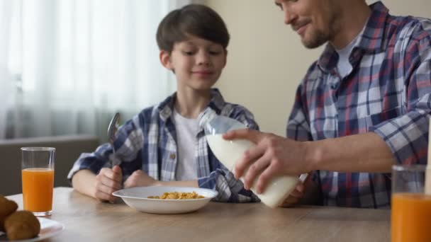 Padre vertiendo leche en el plato con hojuelas de maíz, hijo y padre desayunando — Vídeo de stock