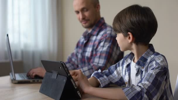 Male dziecko gry na tablet, podczas gdy ojciec pracuje na laptopie w domu, gadżet — Wideo stockowe