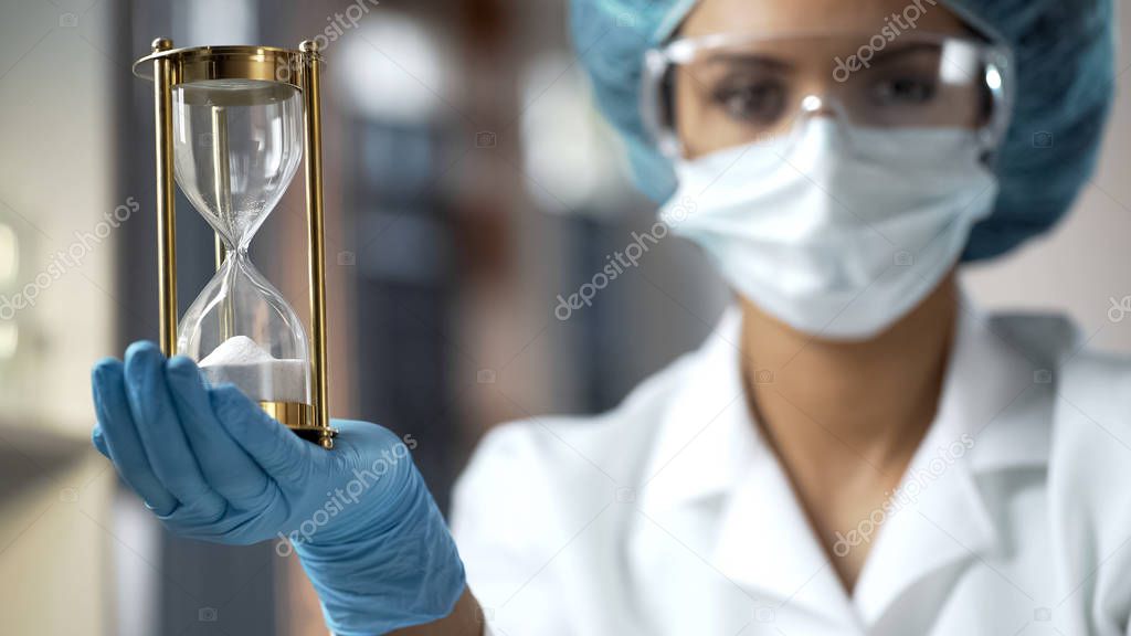 Laboratory worker with hourglass on palm looking camera, limited treatment time