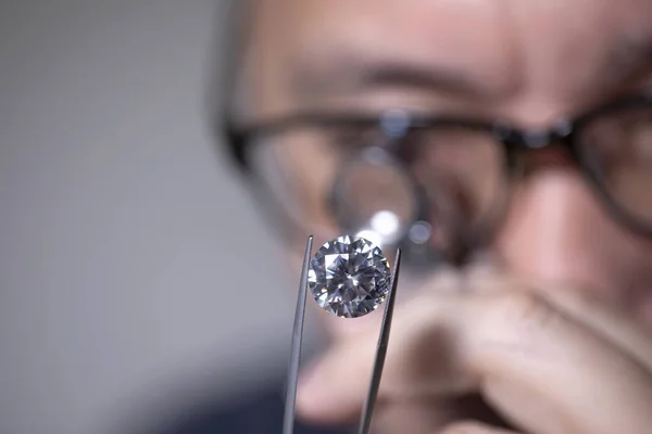 Man jeweller looking through loupe to diamond. Cut and polished diamond. Diamond grading. Diamond trading. Diamond sales. Jeweller holding magnifier. Round diamond. Jeweller on black background