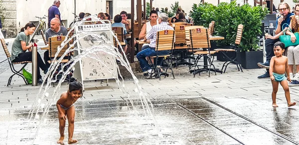 Salzburg / Avusturya 28 Ağustos 2019 - Şehir meydanında fıskiyenin altında oynayan çocuklar ve yaşlılar yaz havasında dışarıda yemek yiyor ve güneş ışığının tadını çıkarıyorlar.