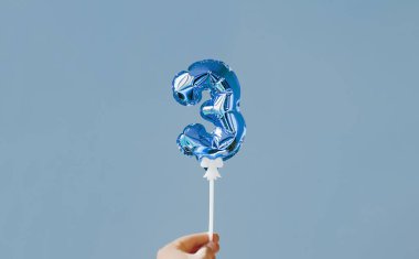 El ele tutuşan mavi folyo helyum 3 numaralı balonu mavi arka planda beyaz bir plastik çubukta şişiriyor. Parlak bayram selamlama düzeni