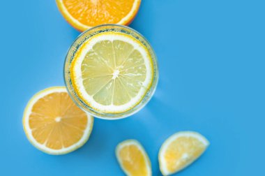 Bardakta yuvarlak bir dilim limon ve mavi arka planda koca bir limon olan yarı saydam gazlı ve köpüklü su bardağı. Sıcakta ferahlatıcı. Sağlıklı yaşam tarzı ve beslenme, üst görünüm