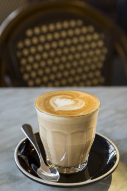 Kahve fincanındaki kahve, sevgi dolu kalp şeklinde sütlü latte sanatı mermer bir masa üzerinde ve kahve dükkanındaki retro sandalye arka planı, dikey görünüm.