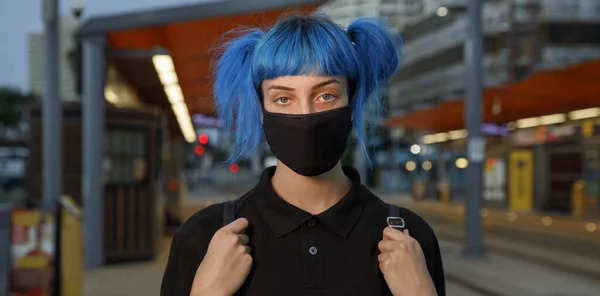 Máscara de la cara médica de moda usada por las mujeres jóvenes con el pelo azul en la parada del tranvía en una calle urbana de la ciudad en el atardecer Fotos de stock libres de derechos