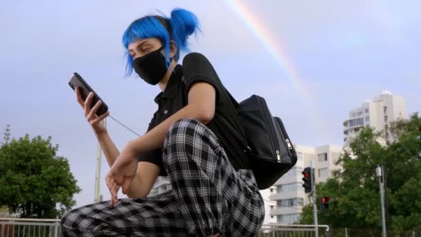 年轻女孩戴着医用口罩 头戴验尸官式的恐惧 穿着蓝色头发的时髦女生 带着彩虹背景的智能手机 新的或现代的时尚观念 — 图库视频影像