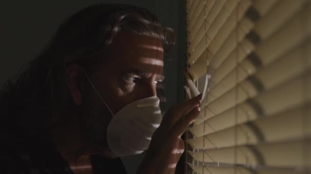 头颈类动物 头戴医用口罩的男性 带着自我隔离的面具 看着窗外的百叶窗外面的家 忧心忡忡的人摘下面具 笑着走在科维德19号检疫病毒大流行后 — 图库视频影像