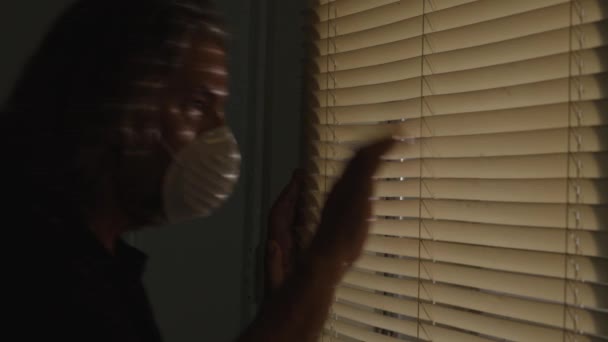 Коронавирус, разочарованный мужчина в медицинской маске в самоизоляции, выглядывающий из оконных жалюзи — стоковое видео