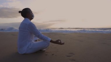 Nilüfer çiçekli tıbbi maske takan adam gün doğumunda sahilde poz verir.