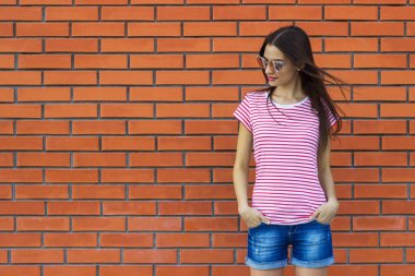 Kırmızı ve beyaz giyen hippi kız t-shirt, moda güneş gözlüğü ve tuğla duvar arka planı, minimalist sokak tarzı poz kot şort çizgili