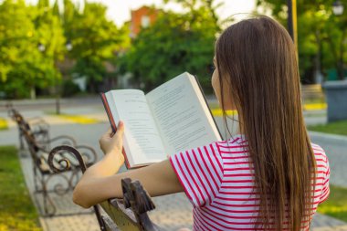 Kırmızı ve Beyaz tişört parkta bankta bir kitap yaz günde zevk çekici kadın. Güzel kız rahatlatır ve parkta bankta bir kitap okur.