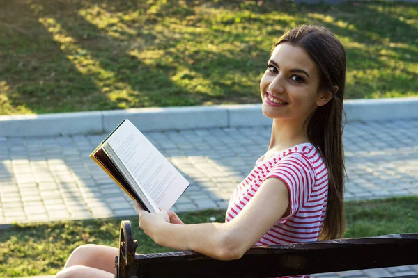 Bir kitap okuma ahşap bir bankta oturan kız gülümsüyor. Pretty genç kız parkta bir kitap okuma sırasında kafa geriye döner. Yan görünüm