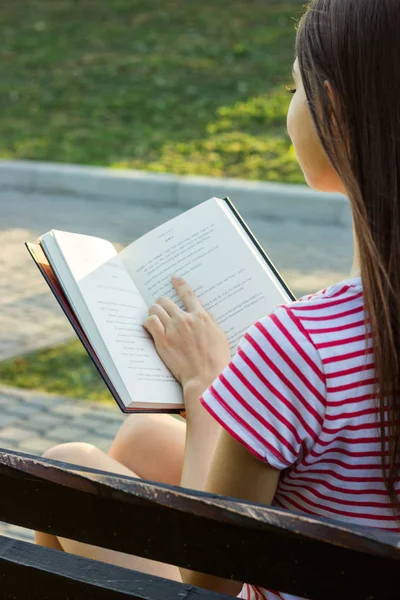 Menina bonita relaxa e lê um livro no banco no parque. Uma mulher mantém um dedo no texto durante a leitura. Visão traseira — Fotografia de Stock
