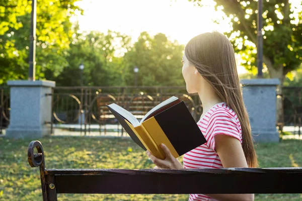 A side view of stunning young girl reading a book in the park in summer sunny day. — Stock Photo, Image