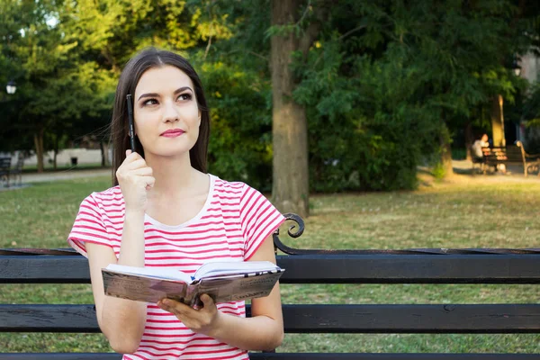 Park ahşap bankta oturmuş ve bir kitap ve kafasını tarafından yerleştirilen bir kalem ile düşünme güzel genç kız