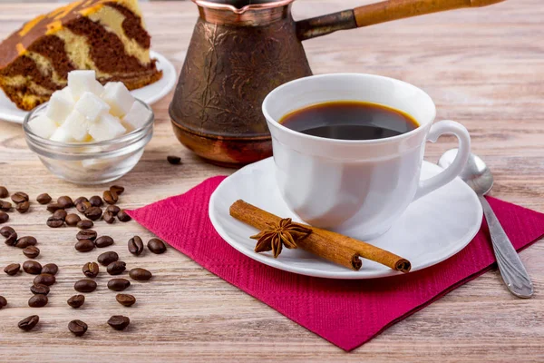 Et stykke sjokoladekake med hvit kopp svart, varm kaffe på tallerken, servert med kaffebønner, hvite sukkerbiter i en bolle, teskje, anis og kanelpinner på mørk trebakgrunn – stockfoto