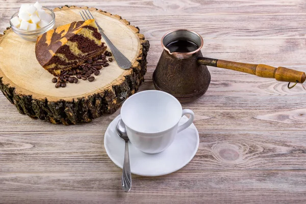 Un delicioso pastel en tocón de madera con una taza de café, tenedor, cuchara de té, granos de café y tazón con terrones de azúcar sobre un fondo de madera — Foto de Stock