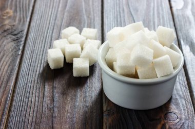 Beyaz ahşap arka plan üzerinde kapta şeker. Seçici odak, yatay. Bir kaç küp şeker bardağı beyaz şeker ile yakındır. Kötü kalori alımı.