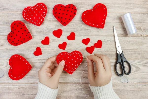 Valentine day theme. Workplace for preparing handmade decorations. Top view of female hands sew felt heart. Packed gifts, tools shabby wooden table