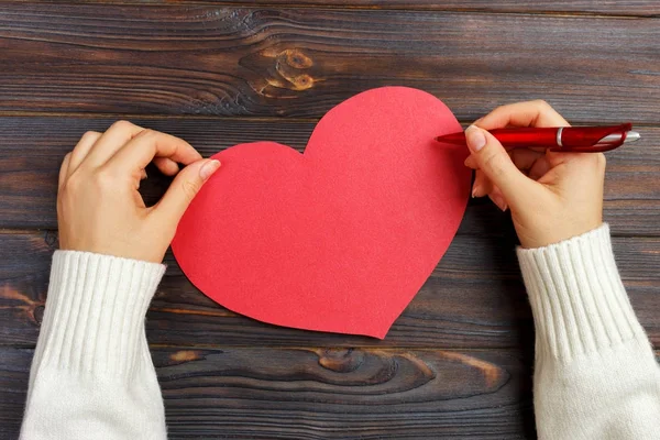 Hand of girl writing love letter on Valentine Day. Handmade red heart postcard. Woman write on postcard for 14 February holiday celebration. Flat lay. Top view