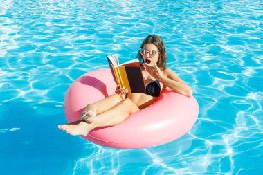 sürpriz kız pembe şişme daire üzerinde bir havuzda bir kitap okuma. Kadın havuzda eğleniyor