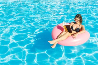 Beautiful woman reading a book on inflatable ring in blue swimming pool clipart