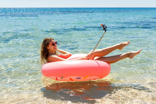 Технологии и концепция людей - улыбающаяся молодая женщина или девочка-подросток делает селфи на надувном пончике со смартфоном на пляже — стоковое фото