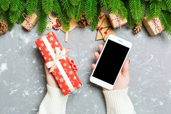 Top view of a woman holding a phone in one hand and a gift in another on cement background. Fir tree and holiday decorations. Christmas holiday concept. Mockup