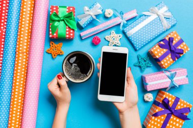 Bir elinde telefon, diğer elinde kahve tutan bir kadının mavi arka plandaki görüntüsü. Noel süslemeleri ve oyuncakları. Yeni yıl tatili konsepti. Model