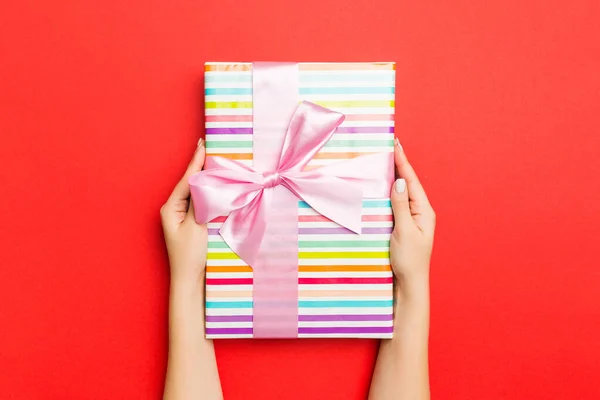 Mãos femininas segurando caixa de presente listrada com fita colorida no fundo vermelho. Conceito de Natal ou outra caixa de presente artesanal de férias, conceito vista superior com espaço de cópia — Fotografia de Stock