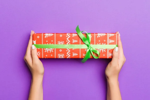 Colocación plana de manos de mujer sosteniendo regalo envuelto y decorado con arco sobre fondo púrpura con espacio de copia. Concepto de Navidad y vacaciones — Foto de Stock