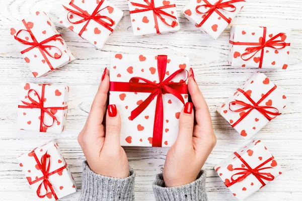 Woman hands give wrapped valentine or other holiday handmade present in paper with red ribbon. Present box, red heart decoration of gift on wooden table, top view with copy space for you design