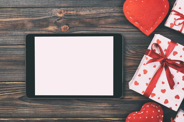 Digital tablet blank screen with gift box and hearts decor on wooden table. Top view. Valentines Day concept background