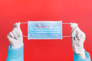 Female hands in medical gloves holding protective mask with pandemic word on it at red background. Health care concept. Coronavirus concept