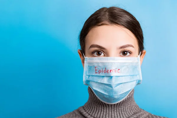 Retrato de jovem mulher usando máscara médica com palavra epidêmica em fundo azul. Proteja a sua saúde. Conceito de coronavírus — Fotografia de Stock