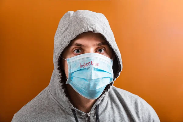 Portrait of young man in hood wearing protective medical mask with infection text at brown background. Coronavirus concept. Healthcare concept — 스톡 사진