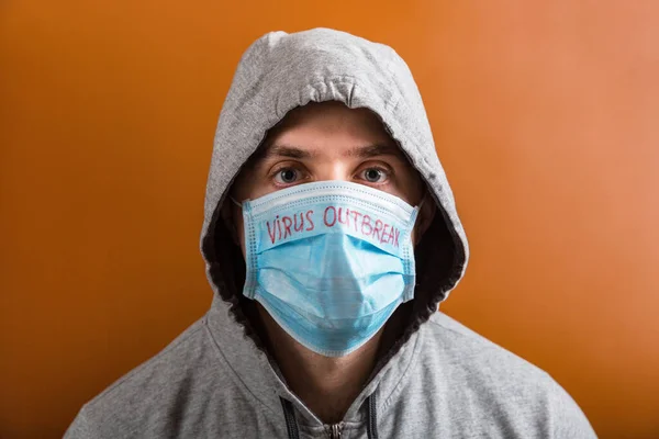 Portrait of young man in hood wearing protective medical mask with virus outbreak text at brown background. Coronavirus concept. Healthcare concept — 스톡 사진