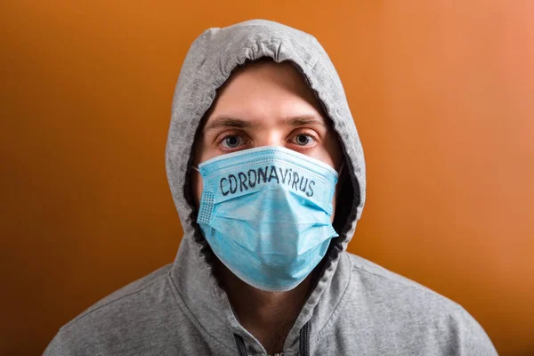 Portrait of young man in hood wearing protective medical mask with coronavirus text at brown background. Coronavirus concept. Healthcare concept — 스톡 사진