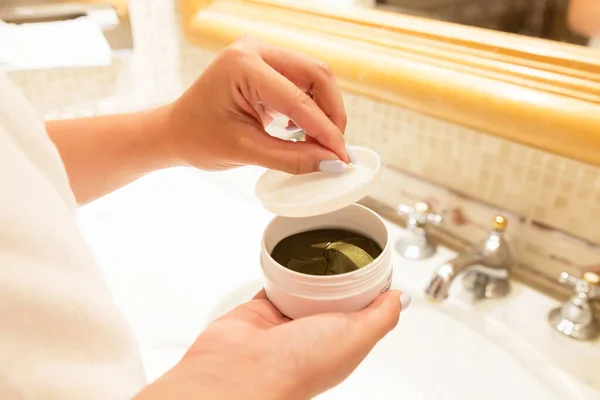 Top view of female hands opening a jar with eye patches in the bathroom. Hydrogel patches against dark circles under eyes.