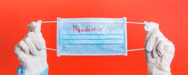 Female hands in medical gloves holding protective mask with pandemic word on it at red background. Health care concept. Coronavirus concept.