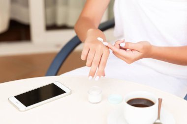Cropped image of female hands applying cream at the hotel terrace. Phone and a cup of coffee on the table. Skincare concept.
