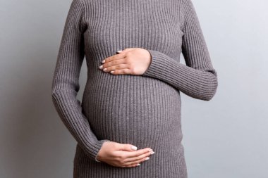 Gri elbiseli hamile bir kadının gri arka planda büyük karnını kucaklarken görüntüsü. Annelik konsepti. Boşluğu kopyala.